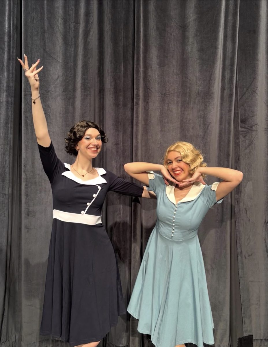 Female student in black dress stands next to female student in blonde wig and blue dress on stage